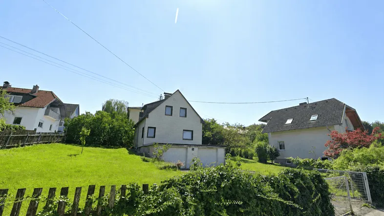 Blick auf ein Einfamilienhaus mit großem Garten und Holzzaun an einem sonnigen Tag.