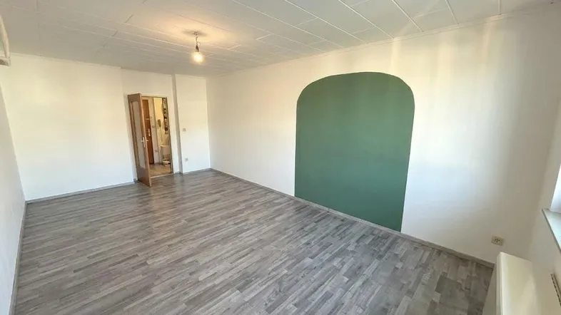 Helles, unmöbliertes Zimmer mit modernem Laminatboden und einer grünen Akzentwand.