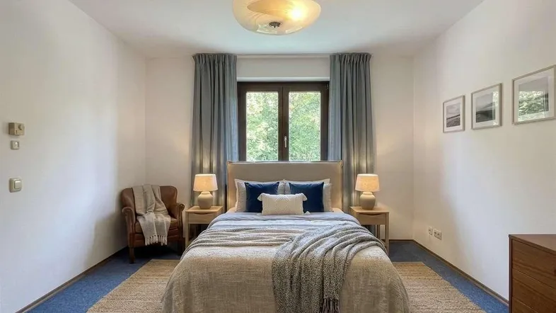 Helles Schlafzimmer mit blauem Teppichboden, beigem Bett und einem Fenster mit Blick ins Grüne.