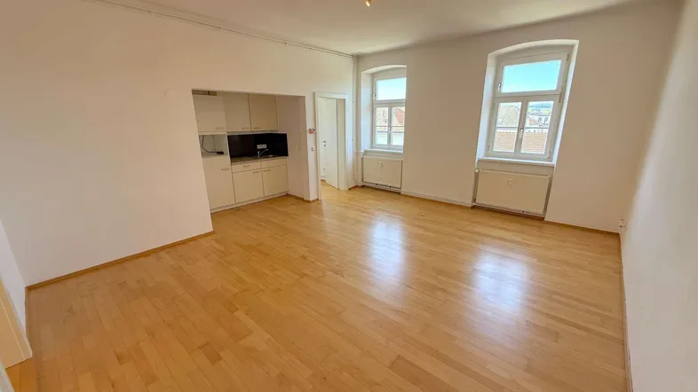 Helles Zimmer mit Holzboden, zwei Fenstern und integrierter Küchenzeile in einem offenen Bereich.
