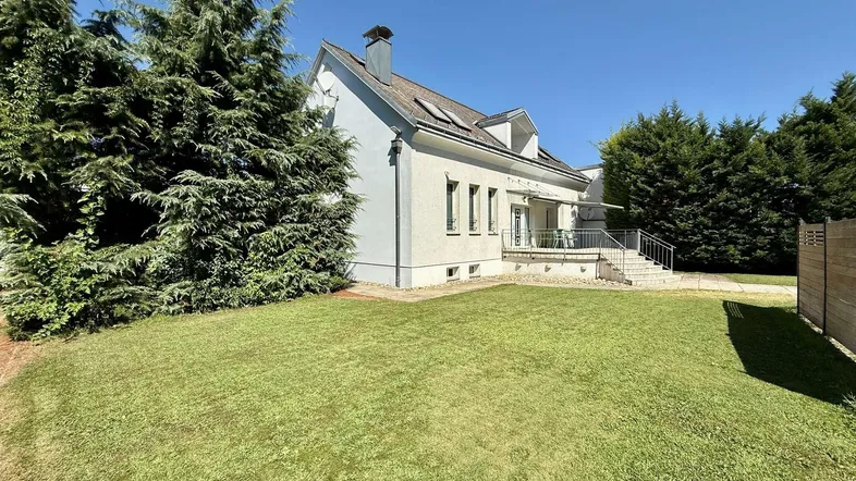 Weißes Einfamilienhaus mit gepflegtem Rasen und hohen Bäumen unter blauem Himmel.