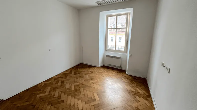 Helles Zimmer mit Fischgrätparkettboden und einem Fenster, das Tageslicht hereinlässt.