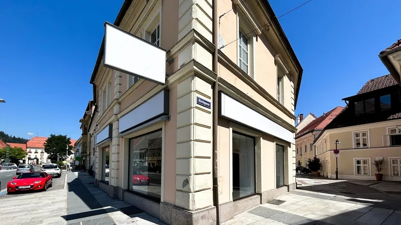 Gebäude an einer Straßenecke mit großen Schaufenstern und leeren Werbetafeln unter blauem Himmel.