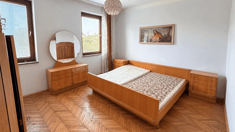 Helles Schlafzimmer mit Holzmöbeln, Fischgrätparkett und zwei Fenstern, die viel Licht hereinlassen.