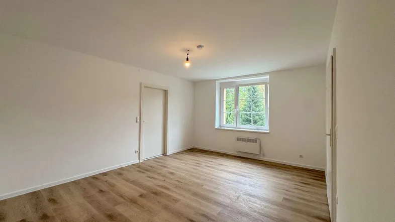 Helles, leeres Zimmer mit Laminatboden und einem Fenster, das viel Tageslicht hereinlässt.