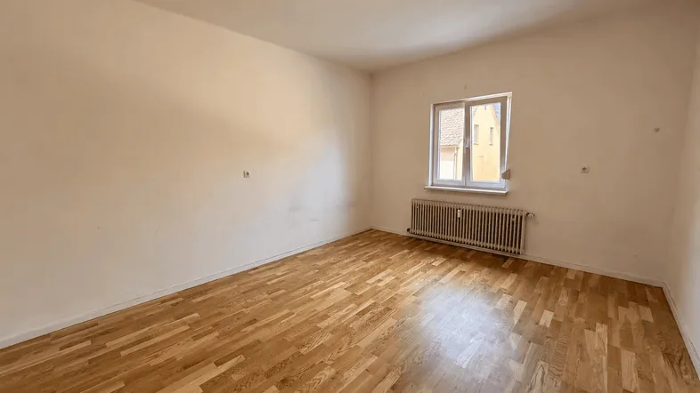 Helles, leeres Zimmer mit Holzparkettboden und einem Fenster, das Tageslicht hereinlässt.