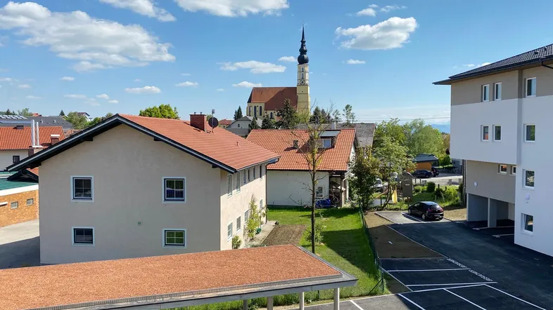 Blick auf die Wohnanlage mit Parkplätzen, umliegenden Gebäuden und einer Kirche im Hintergrund unter blauem Himmel.