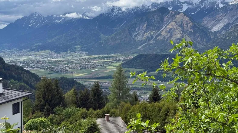 Panoramablick auf die umliegende Berglandschaft und das Tal mit vereinzelten Häusern unter bewölktem Himmel.