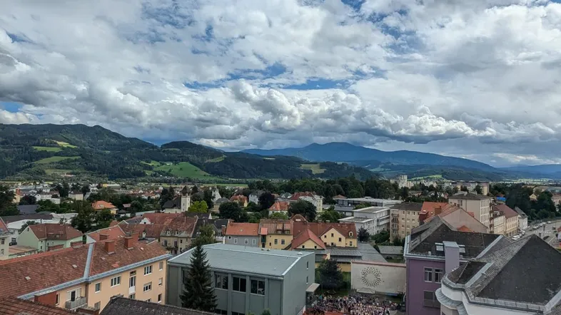 Panoramablick über die Stadt mit roten Dächern und majestätischen Bergen im Hintergrund unter bewölktem Himmel.