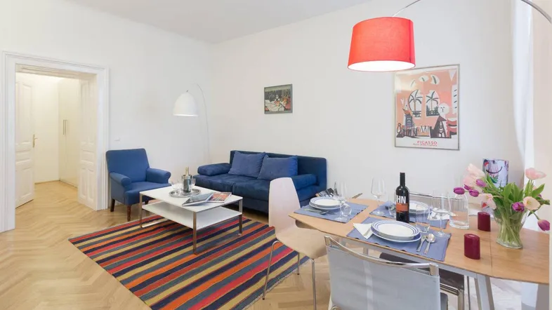 Helles Wohn- und Esszimmer mit Parkettboden, blauer Couch und farbenfrohem Teppich.