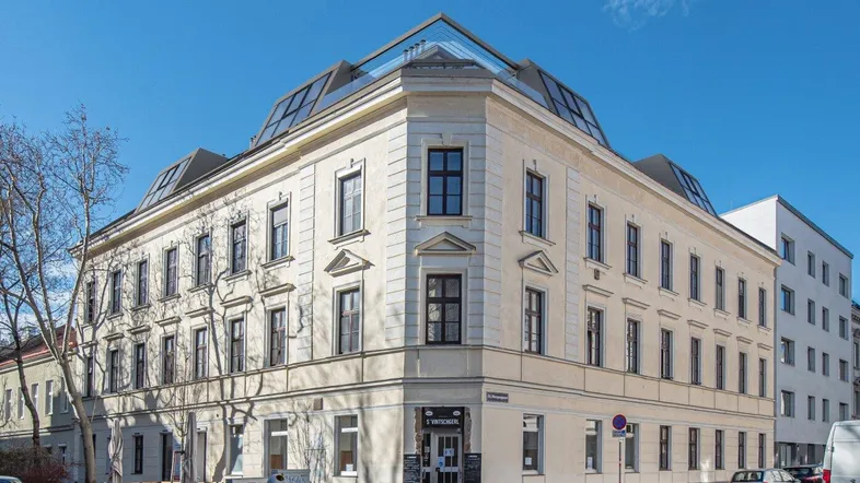Fassade eines Eckgebäudes mit klassischer Architektur und modernen Dachaufbauten unter blauem Himmel.