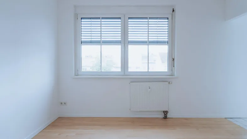 Helles Zimmer mit Holzfußboden und Fenster mit Jalousien, bereit zur individuellen Gestaltung.