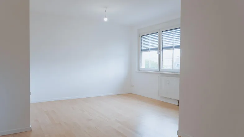 Heller, leerer Raum mit Holzboden und großem Fenster mit Jalousien, viel Tageslicht.