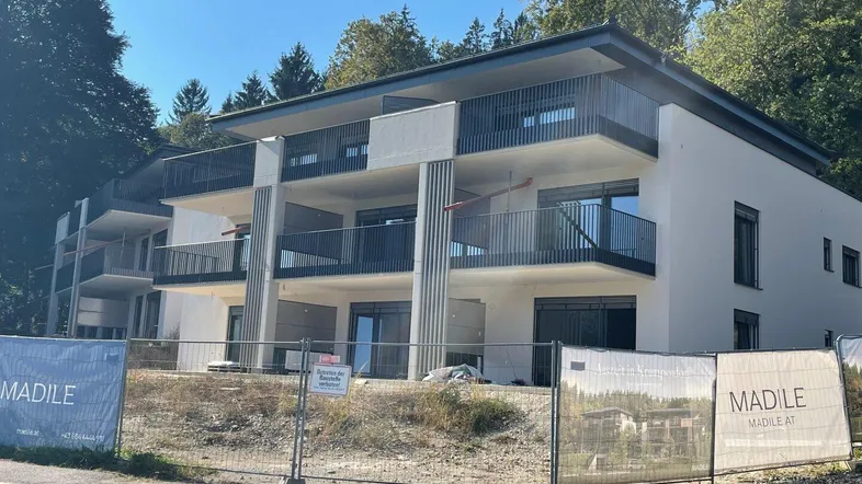 Modernes Mehrfamilienhaus im Bau mit mehreren Balkonen und Flachdach, umgeben von viel Grün.