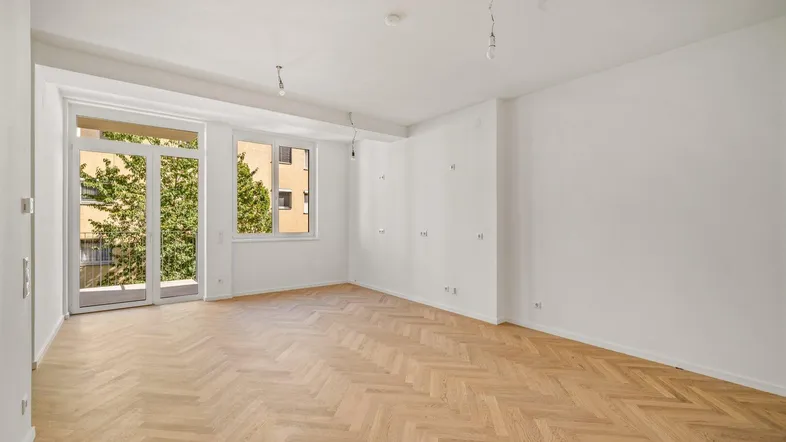 Helles, leeres Wohnzimmer mit Fischgrätparkett und Zugang zu einem Balkon, umgeben von grünen Bäumen.