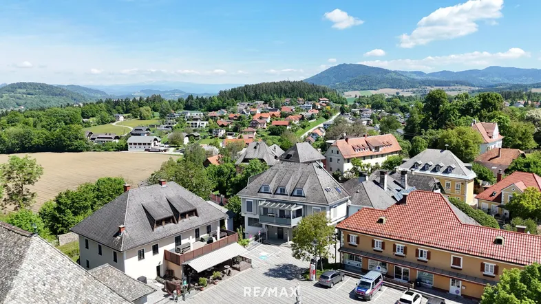Luftaufnahme eines charmanten Dorfes mit traditionellen Gebäuden, umgeben von grünen Hügeln und Feldern unter blauem Himmel.