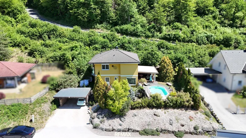 Vogelperspektive eines gelben Einfamilienhauses mit Pool und Carport, umgeben von üppiger Natur.