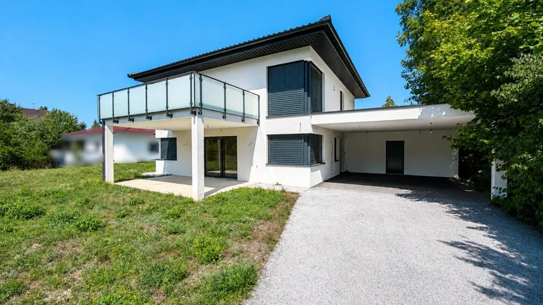 Modernes Einfamilienhaus mit Balkon, Carport und großem Gartenbereich unter blauem Himmel.