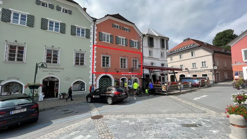 Historisches Gebäude an einem belebten Stadtplatz mit traditioneller Architektur und Geschäften im Erdgeschoss.