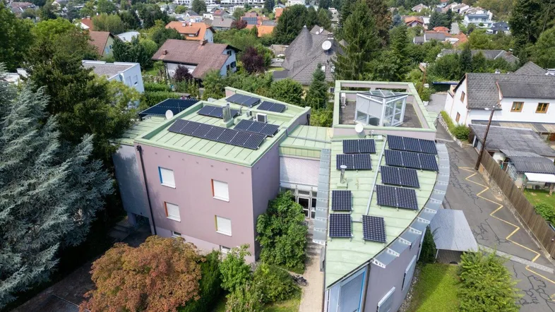 Luftaufnahme eines modernen Gebäudes mit Solarpaneelen auf dem Dach, umgeben von viel Grün und Wohnbebauung.