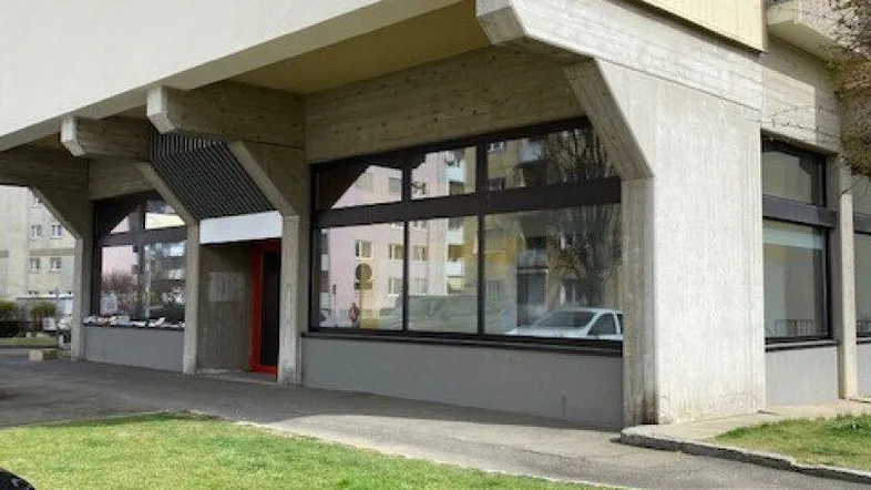 Ansicht der Geschäftsfassade im Erdgeschoss mit großen Schaufenstern und Grünfläche davor.