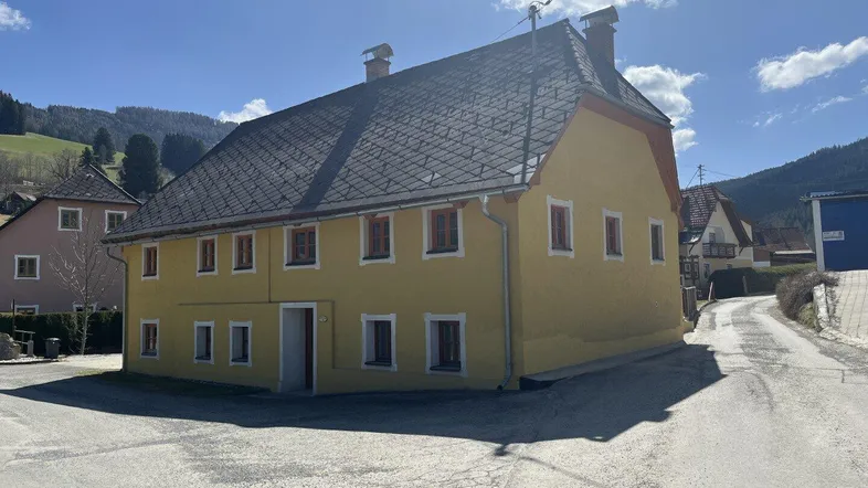 Traditionelles gelbes Haus mit dunklem Dach und mehreren Fenstern in ländlicher Umgebung.