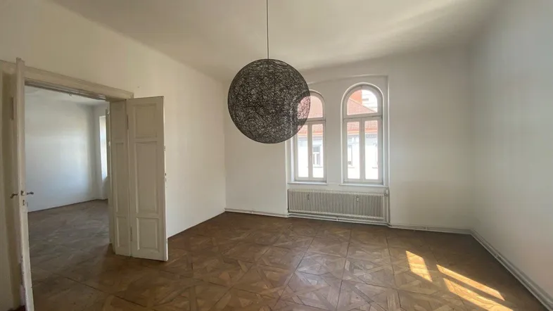 Geräumiges Wohnzimmer mit Fischgrätparkett, hohen Decken und großen Rundbogenfenstern, die viel Tageslicht hereinlassen.