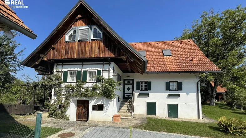 Traditionelles Landhaus mit weißer Fassade, Holzgiebel und rotem Ziegeldach unter blauem Himmel.