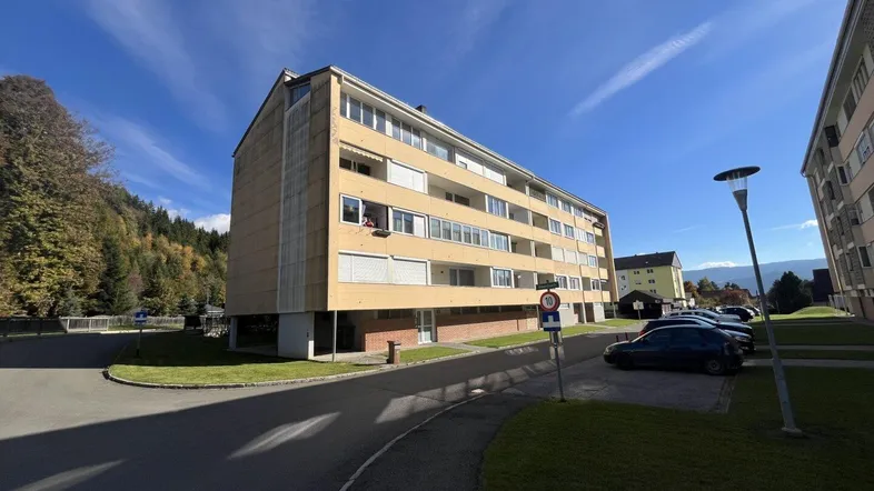 Mehrfamilienhaus mit mehreren Etagen und Balkonen in einer grünen Umgebung unter blauem Himmel.