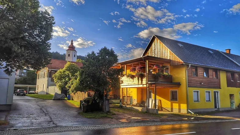 Sonnenbeschienenes Gebäude mit gelber Fassade und Holzbalkon in ländlicher Umgebung bei Sonnenuntergang.