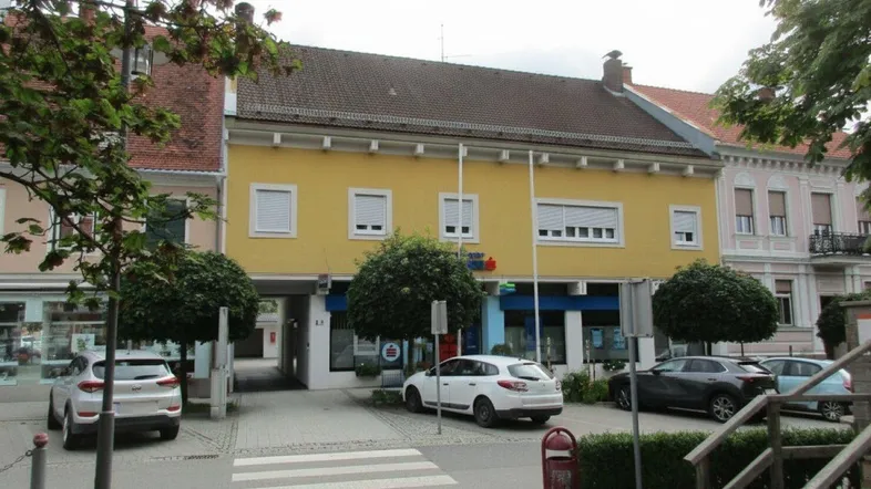 Gelbes Gebäude mit traditioneller Architektur, weißen Fensterläden und einem Durchgang zur Straße. Mehrere Autos sind vor dem Gebäude geparkt.