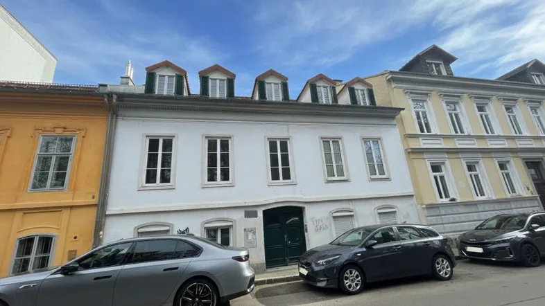 Historisches Gebäude mit weißer Fassade, grünen Fensterläden und Mansarddach in städtischer Umgebung.