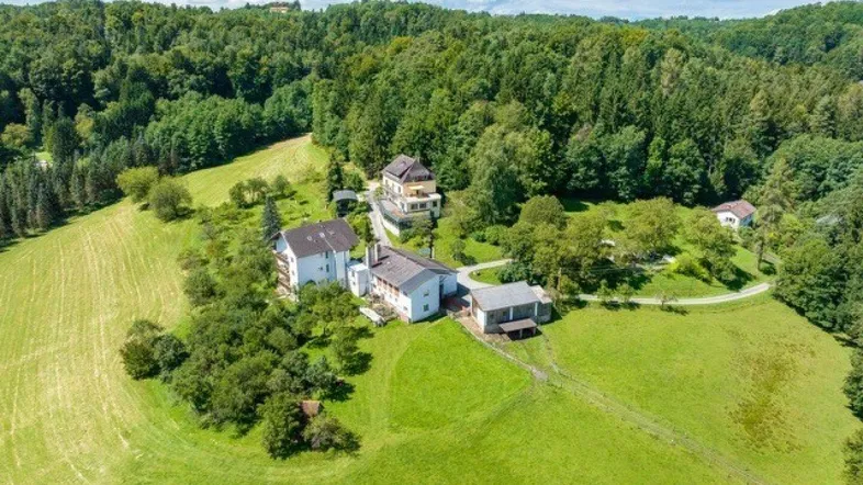 Luftaufnahme eines Anwesens mit mehreren Gebäuden, umgeben von grünen Wiesen und dichtem Wald.