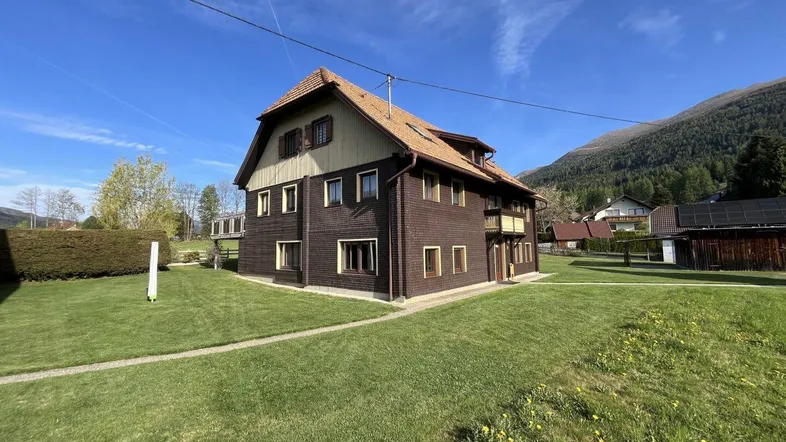 Traditionelles Haus mit Holzfassade und Satteldach, umgeben von einer grünen Wiese und Bergen.