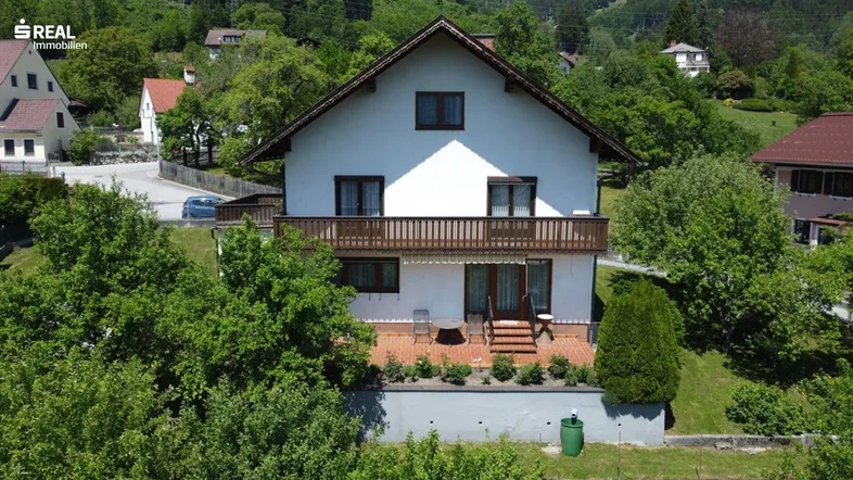 Freistehendes Einfamilienhaus mit Balkon und großer Terrasse, umgeben von viel Grün und Bergen im Hintergrund.