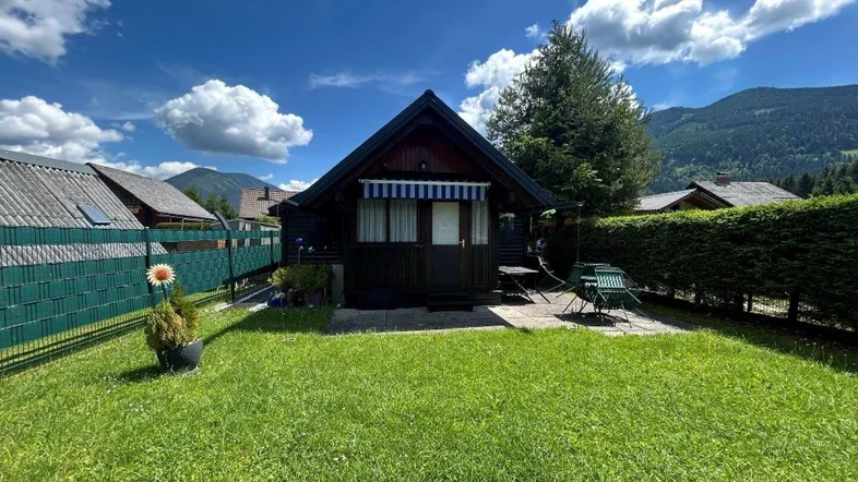 Kleines Holzhaus mit gepflegtem Rasen und Sitzbereich im Freien, umgeben von grüner Natur und Bergen.