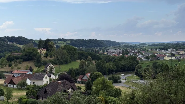 Panoramablick auf ein idyllisches Dorf in einem Tal, umgeben von grünen Hügeln und Wäldern.