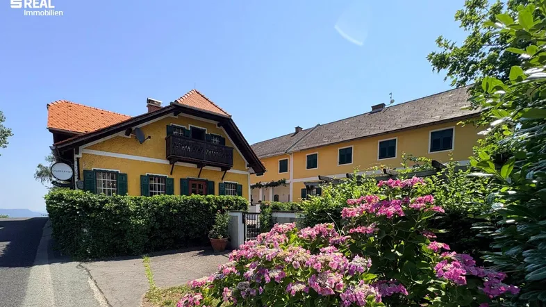 Gelbes Gebäude mit rotem Ziegeldach, grünen Fensterläden und einem kleinen Holzbalkon, umgeben von üppiger Vegetation.