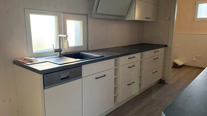 Helle Küche mit Spülmaschine, Spüle unter dem Fenster und Dunstabzugshaube über dem Kochfeld.