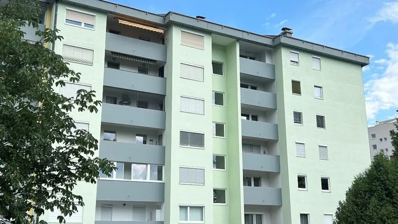 Mehrstöckiges Wohngebäude mit hellgrüner Fassade, Balkonen und Rollläden unter blauem Himmel.