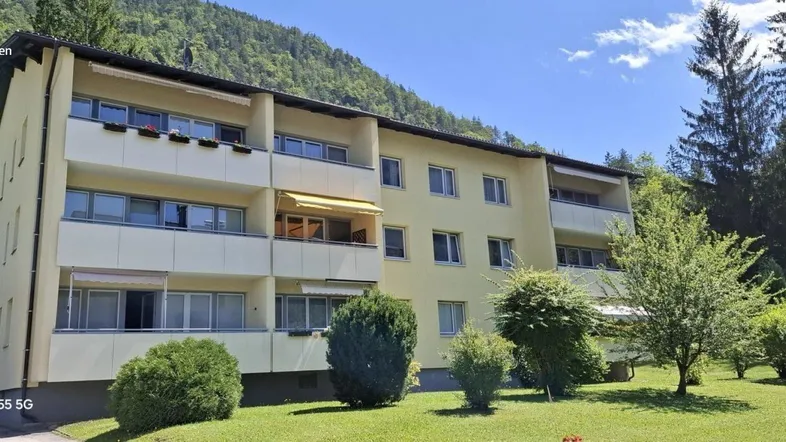 Mehrfamilienhaus mit mehreren Balkonen und grüner Umgebung vor einer Bergkulisse.