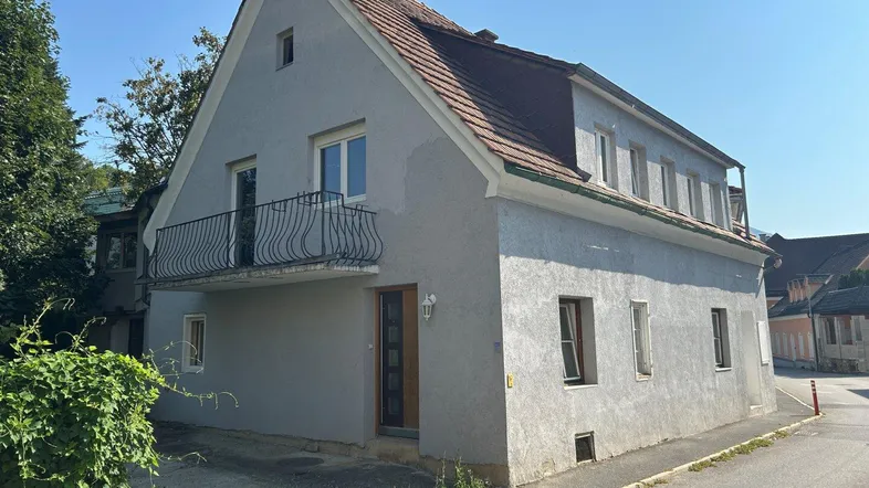 Graues Haus mit Satteldach und kleinem Balkon an der Vorderseite, umgeben von Grün.