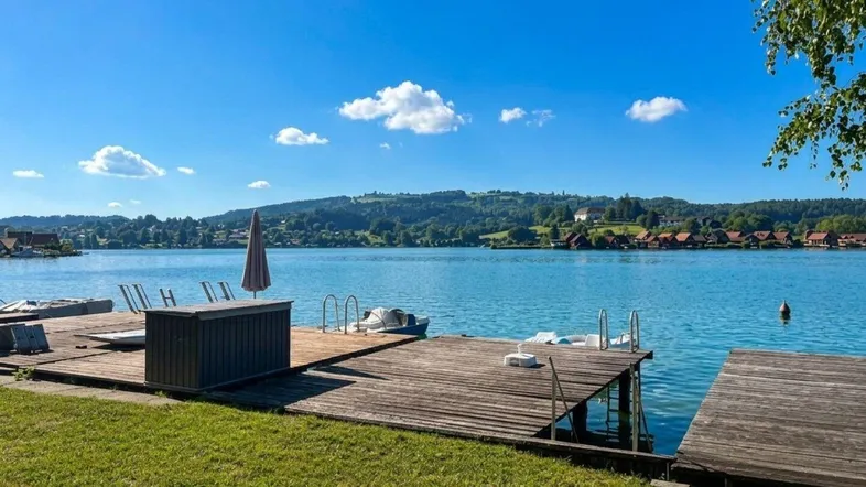 Idyllischer Blick auf den See mit privatem Holzsteg und Liegeflächen unter blauem Himmel.