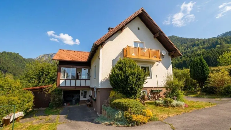 Traditionelles Einfamilienhaus mit Satteldach und Balkon, umgeben von grüner Natur und Bergen.