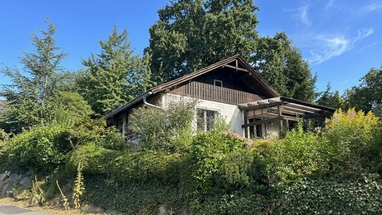 Ländliches Haus mit Satteldach, umgeben von üppiger Vegetation und hohen Bäumen.