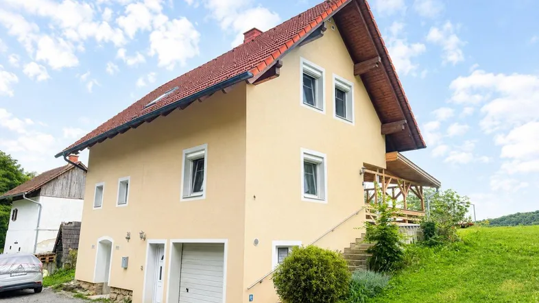 Einfamilienhaus mit gelber Fassade, rotem Ziegeldach und überdachter Holzloggia in ländlicher Umgebung.