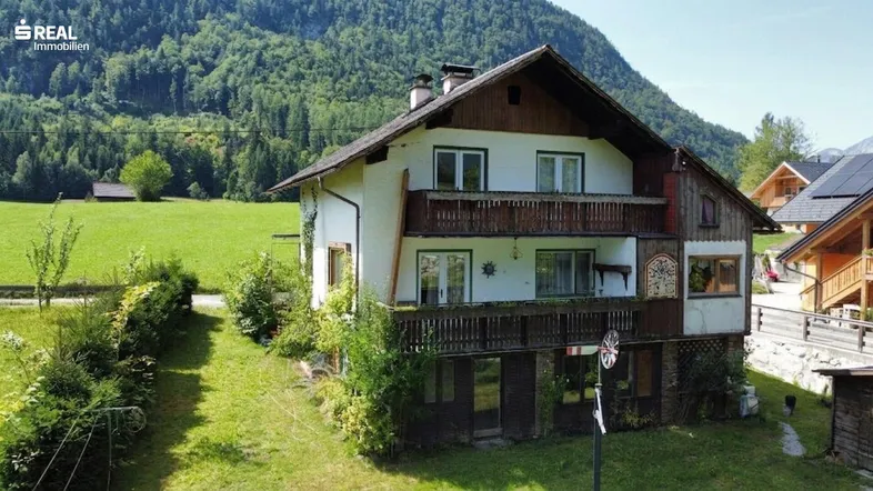 Traditionelles Haus mit Balkonen und Bergblick, umgeben von grünen Wiesen und Wäldern.