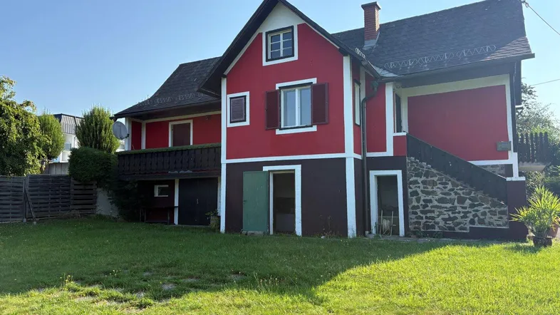 Rotes Haus mit braunem Sockel, grünem Garten und Balkon unter blauem Himmel.
