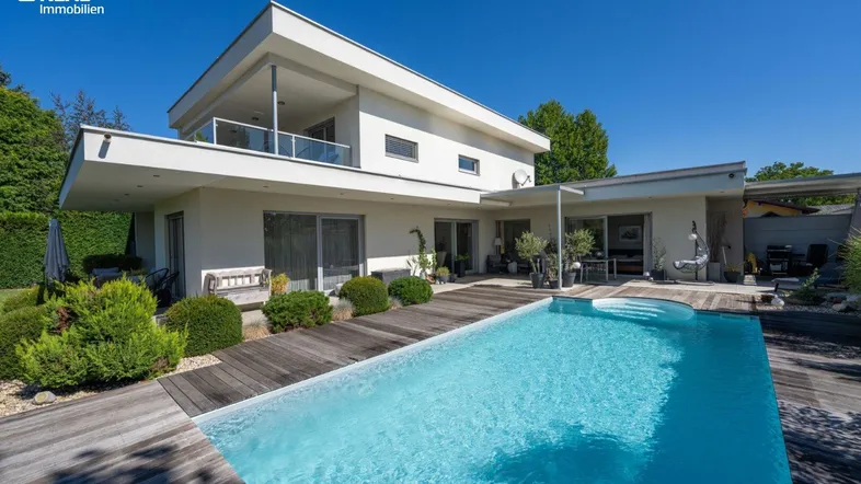 Modernes Haus mit großem Pool und Holzdeck, umgeben von gepflegtem Garten unter blauem Himmel.