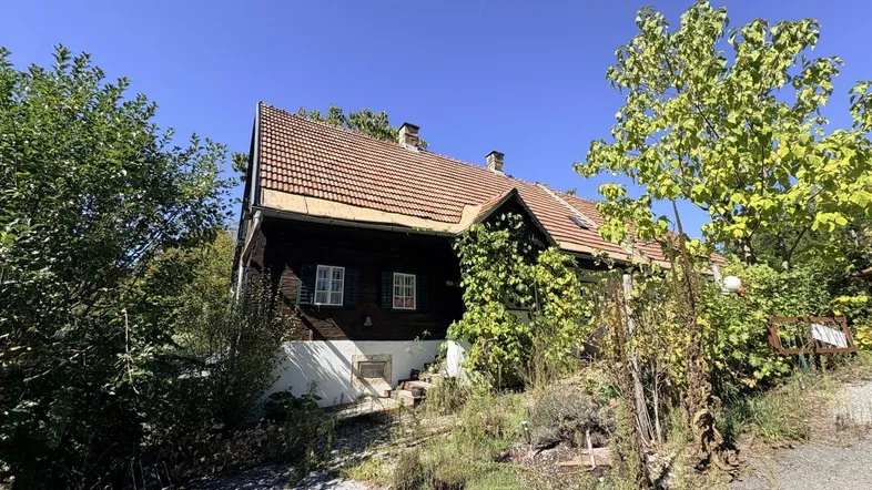 Traditionelles Haus mit Holzelementen und Ziegeldach, umgeben von üppigem Grün unter blauem Himmel.